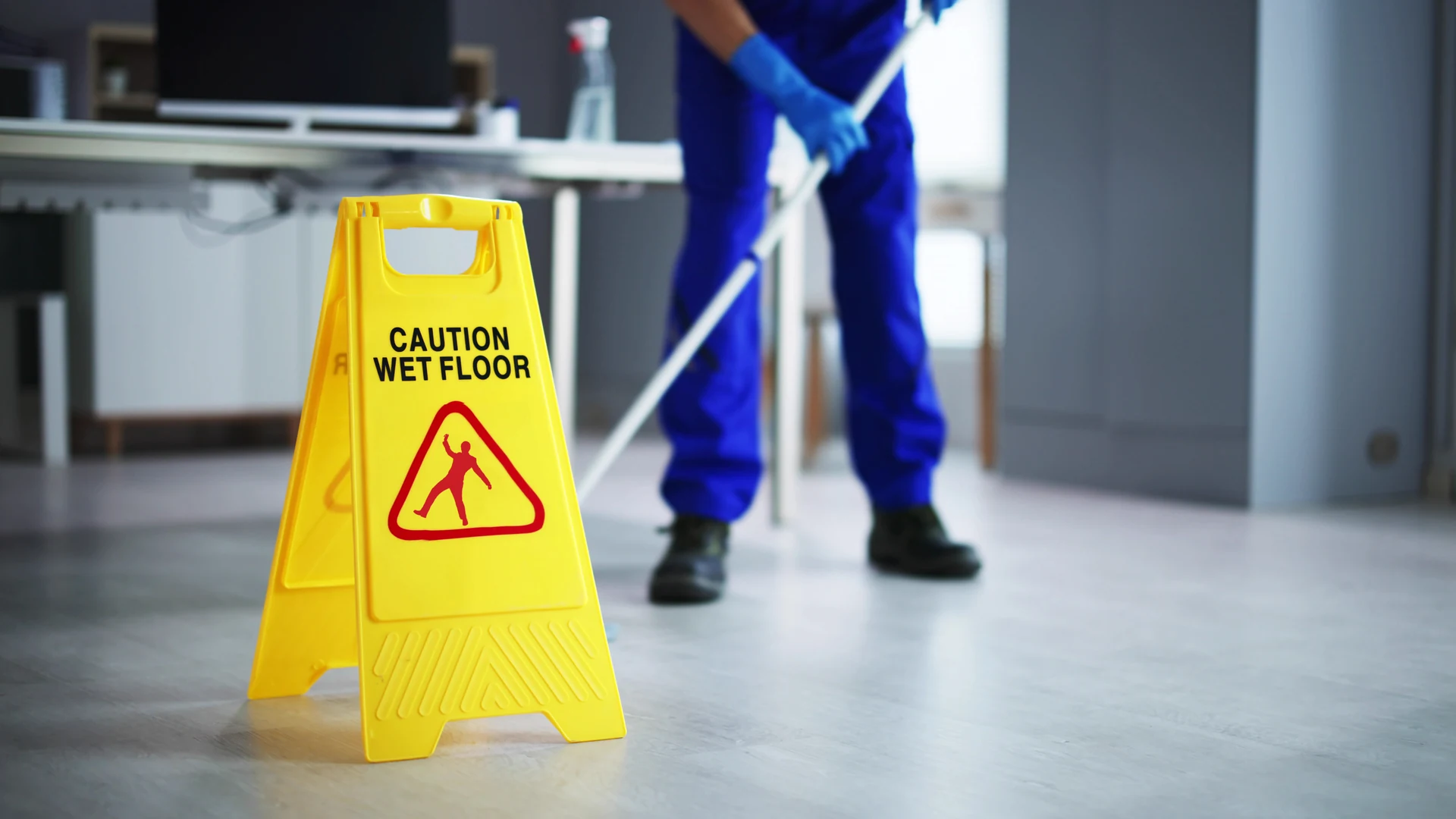 Yellow caution sign on floor with text 'Caution Wet Floor' and a slipping person icon, janitor mopping floor in background.